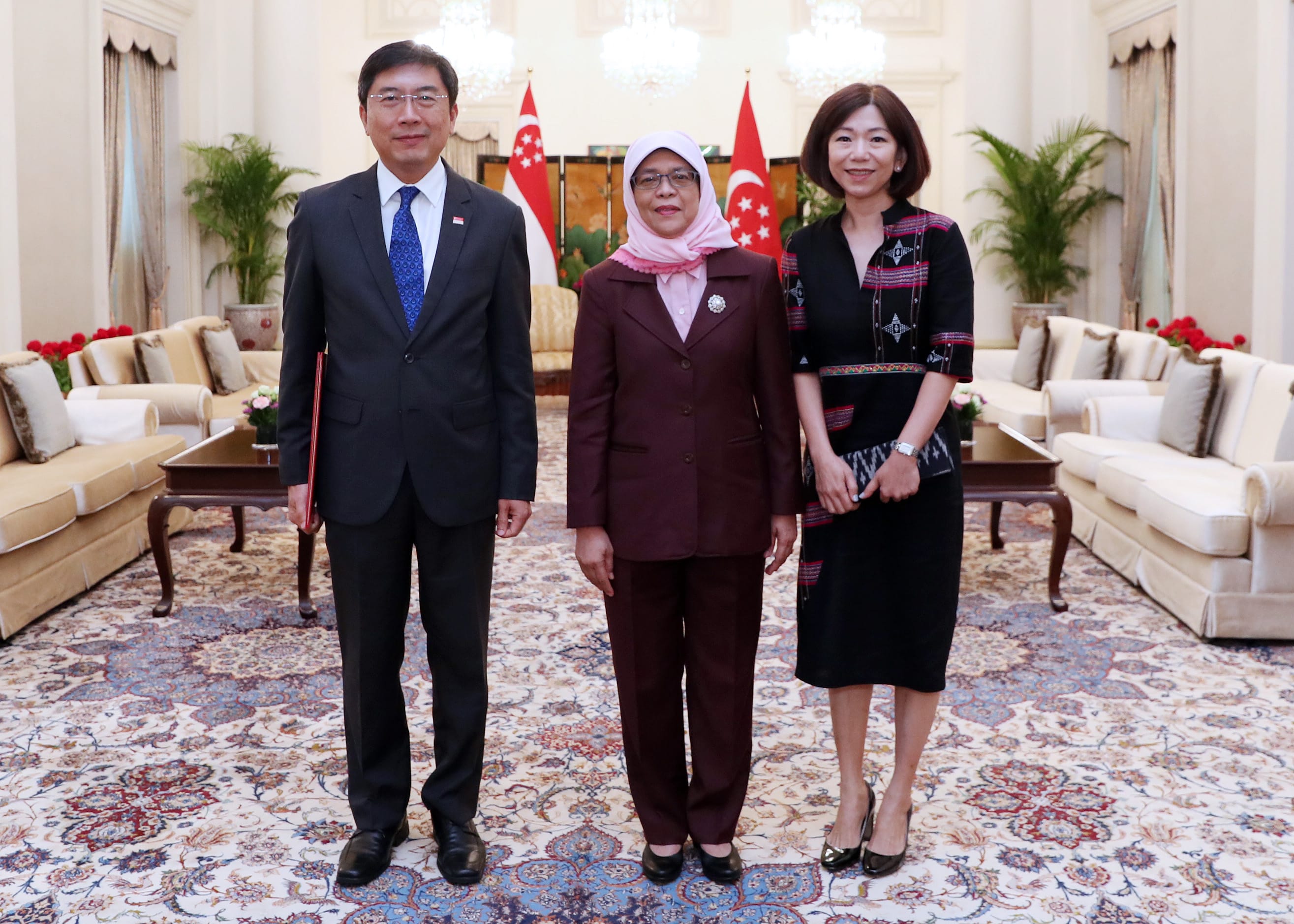 Three people standing indoors, with Singapore flags in the background.